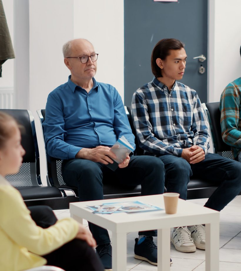 Diverse group of people sitting together in hospital waiting room, preparing to meet with general practitioner at medical clinic. Different patients waiting to attend appointment. Tripod shot.